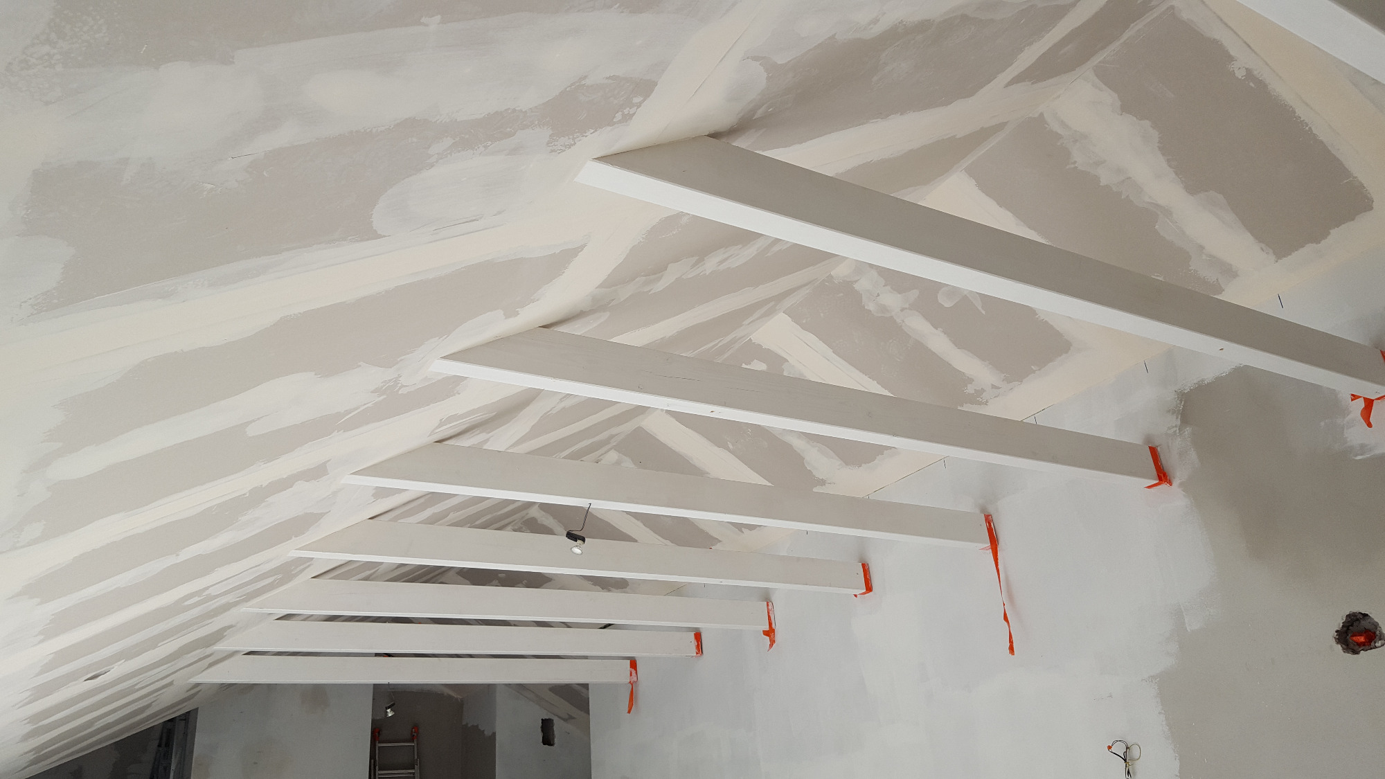 Skośny sufit z płyt kartonowo-gipsowych w trakcie szpachlowania, widoczne białe drewniane belki konstrukcyjne i pomarańczowe elementy mocujące, ślady szpachli na ścianach.
