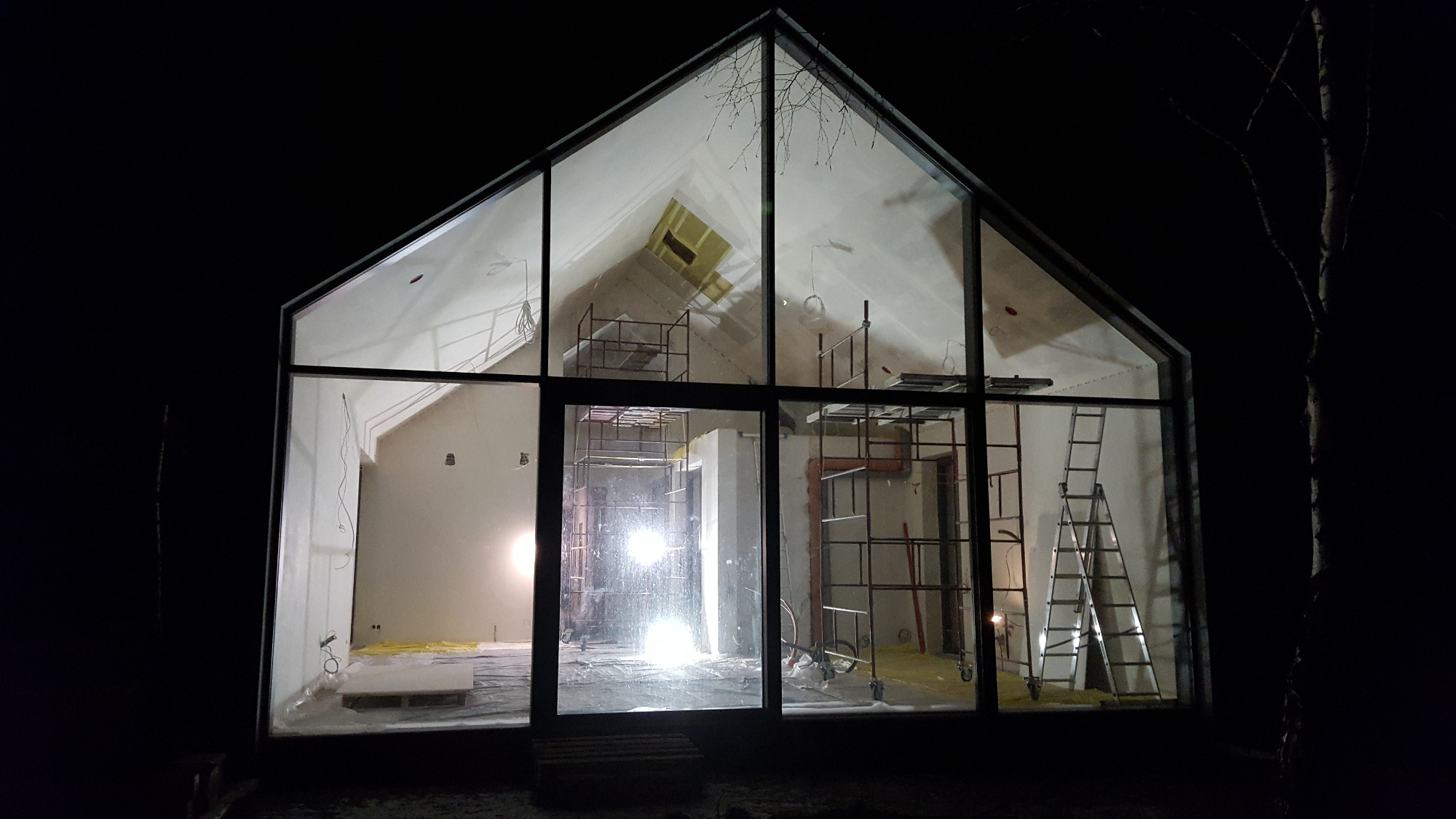 Wnętrze domu w stanie surowym, widoczne przez duże okno z czarnymi ramami, rusztowania i drabina wewnątrz, nocne oświetlenie zewnętrzne.