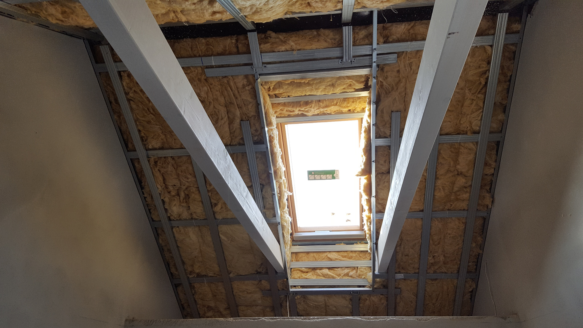 Montaż izolacji z wełny mineralnej na poddaszu, widoczne profile stalowe i drewniane belki, okno dachowe w tle.