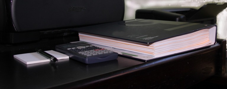 Kalkulator, notatnik z długopisem i segregator z dokumentami na biurku, w tle drukarka.