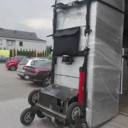 Wózek transportowy z poziomnicą przewozi zapakowany w folię stretch ładunek wewnątrz pojazdu, na zewnątrz widoczny parking z samochodami i budynek.