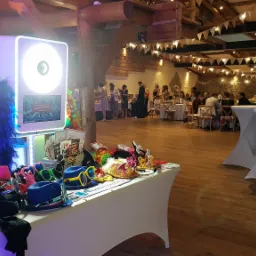 Przygotowana do użycia, oświetlona kabina fotograficzna z rekwizytami na stole w sali balowej, w tle goście weselni przy stołach i girlandy świetlne.