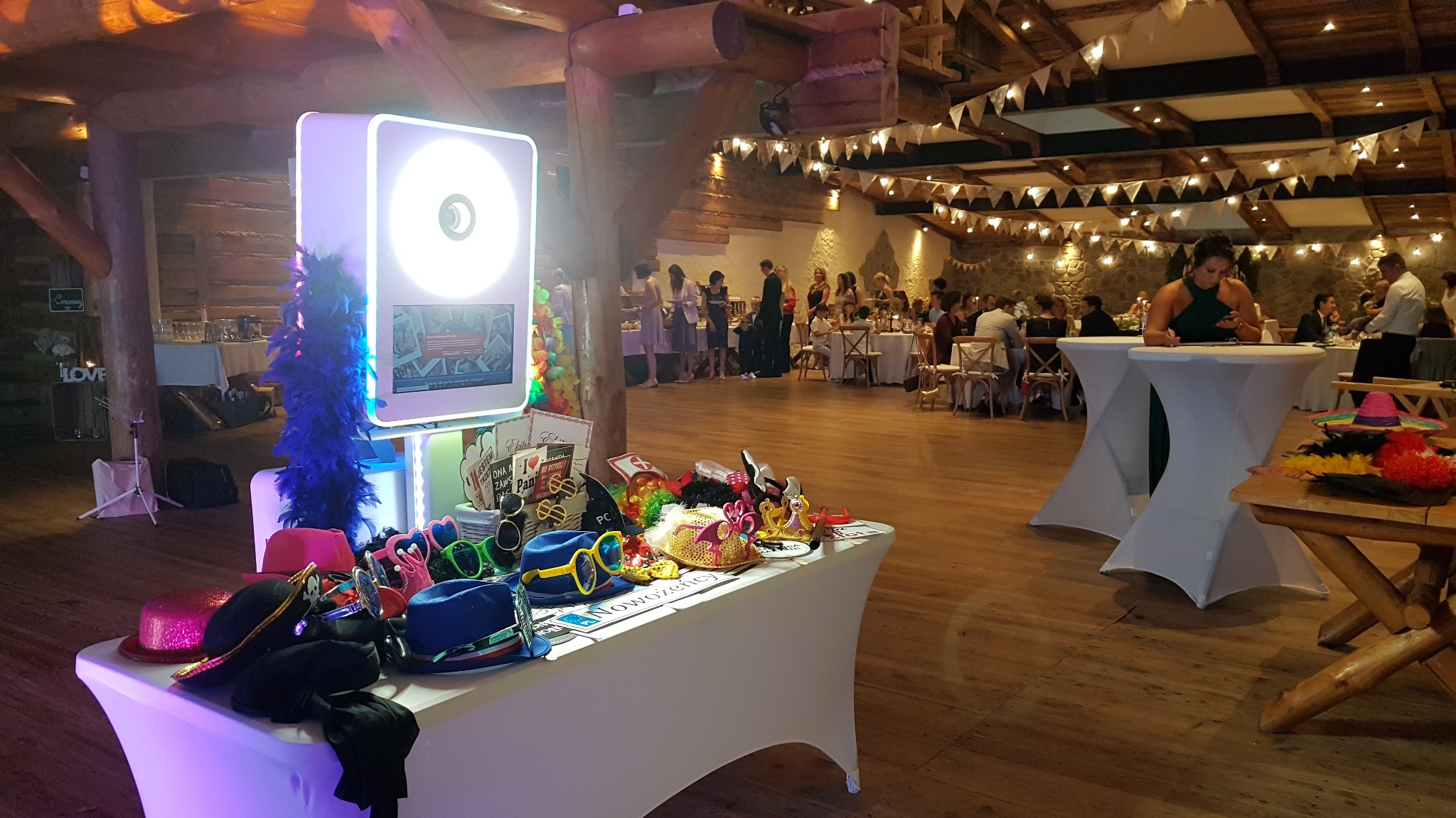 Przygotowana do użycia, oświetlona kabina fotograficzna z rekwizytami na stole w sali balowej, w tle goście weselni przy stołach i girlandy świetlne.