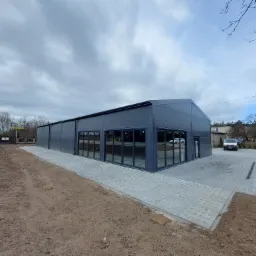 Nowoczesna hala przemysłowa w odcieniach szarości z przeszkloną fasadą, otoczona kostką brukową i przygotowanym terenem pod zieleń, widok z zewnątrz w pochmurny dzień.