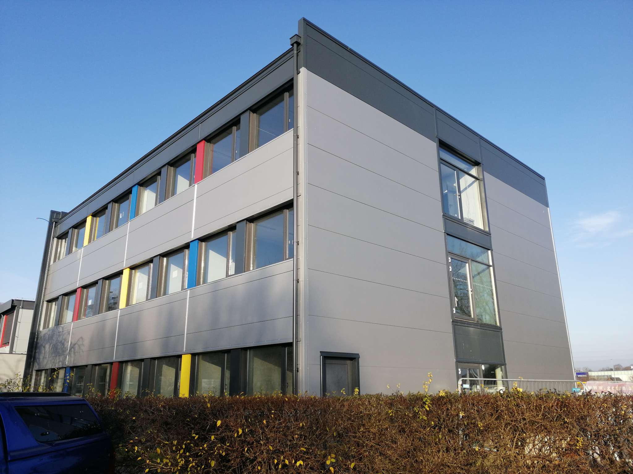 Nowoczesny budynek biurowy z szarych płyt warstwowych, z kolorowymi akcentami wokół okien (czerwony, żółty, niebieski), otoczony niskim żywopłotem i z zaparkowanym niebieskim samochodem w rogu kadru.