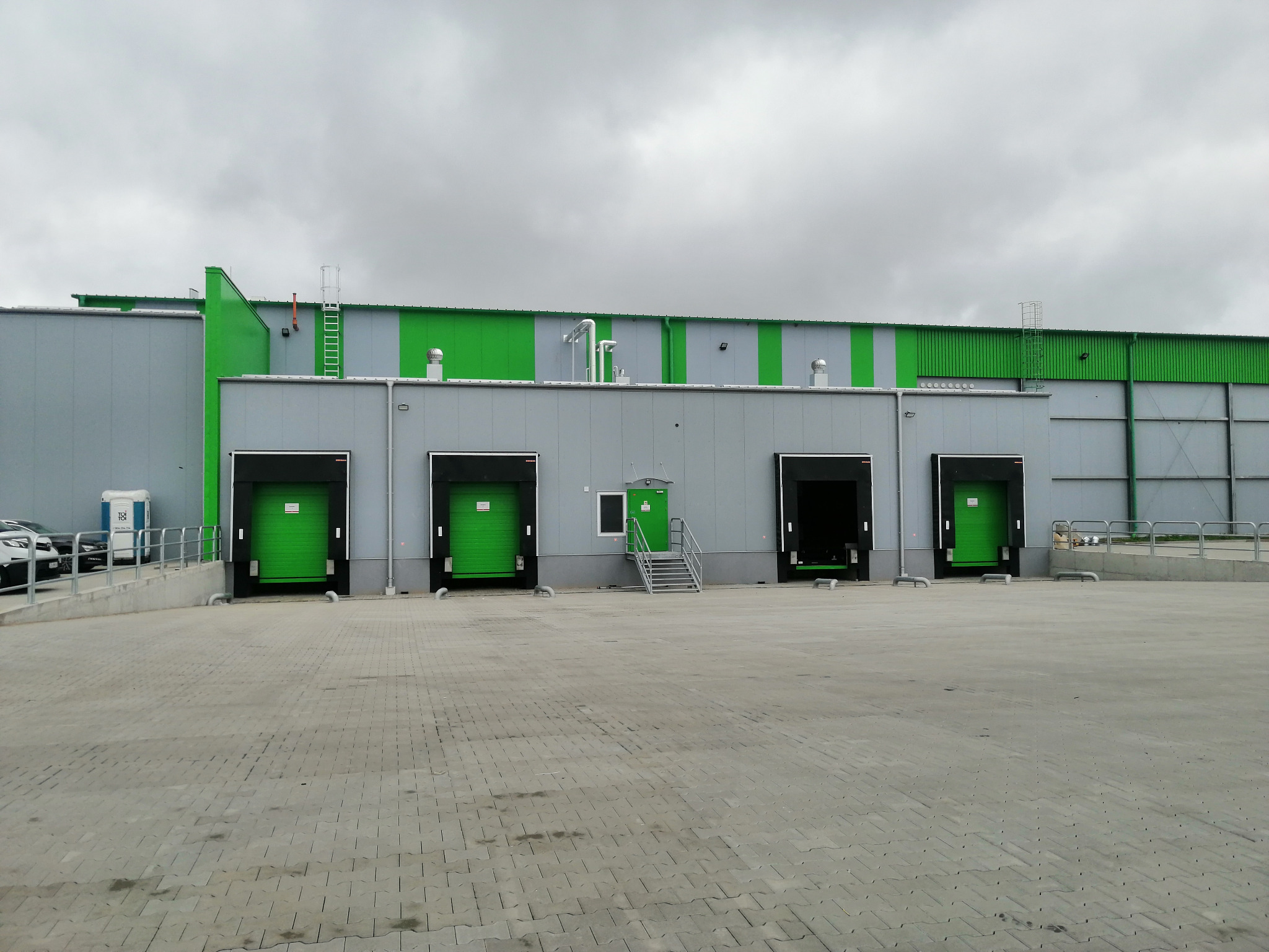 Zewnętrzny widok hali magazynowej z czterema zielonymi bramami segmentowymi, szarą elewacją z zielonymi akcentami, betonowym placem manewrowym i pochmurnym niebem w tle.