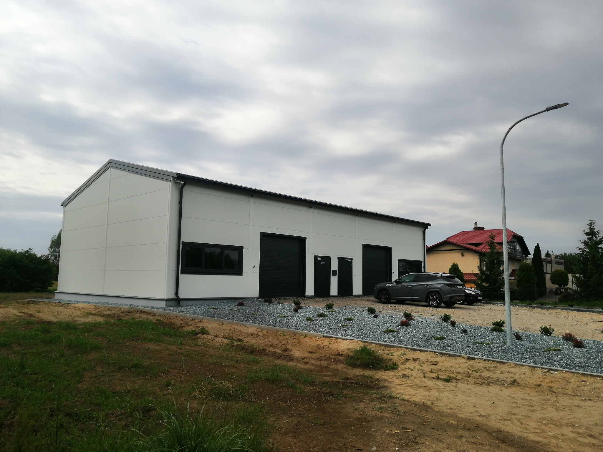 Nowoczesna hala przemysłowa z białych płyt warstwowych, z trzema bramami garażowymi i oknem, obok zaparkowany szary samochód osobowy, teren wokół wysypany ozdobnym żwirem.