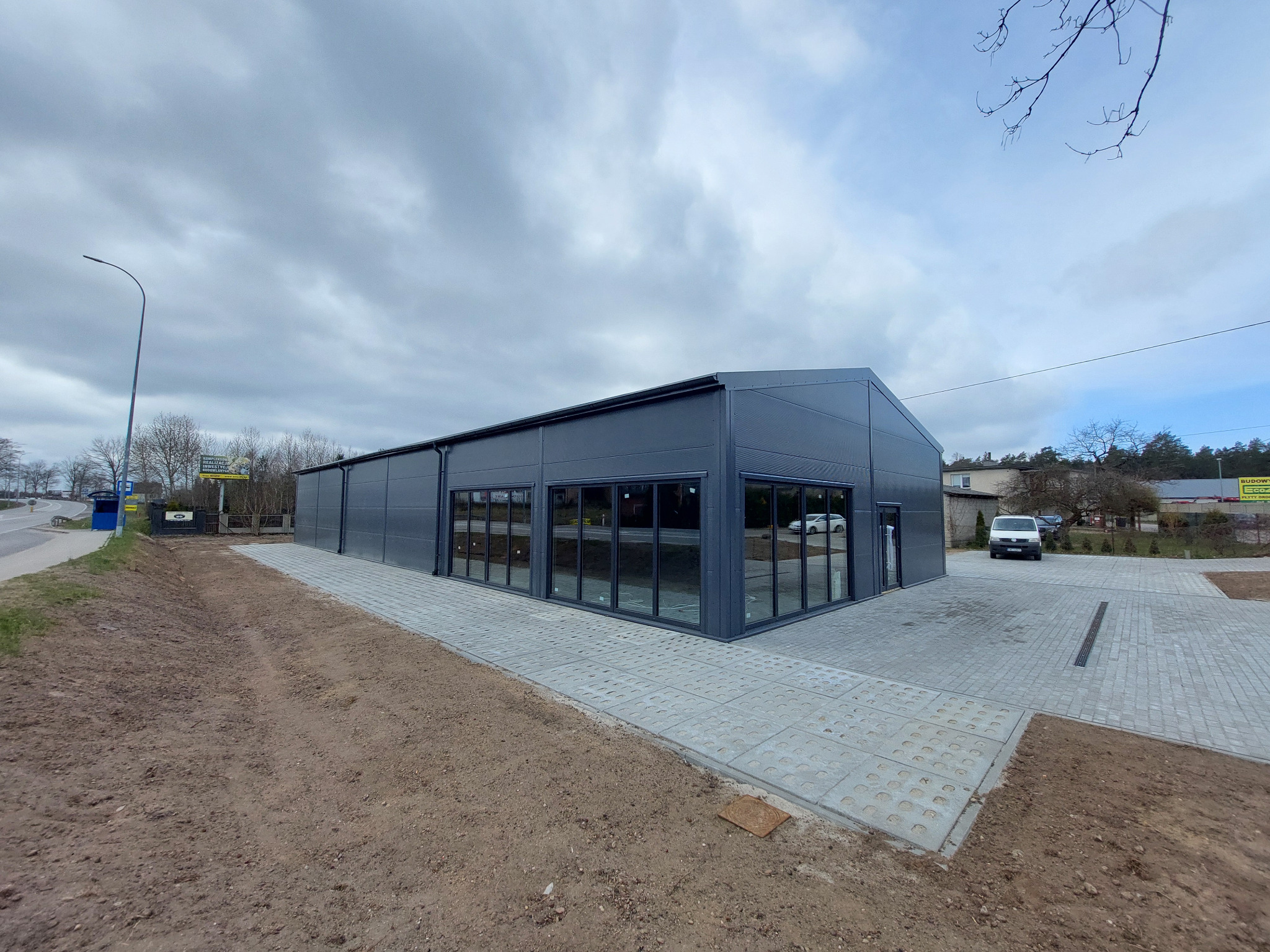 Nowoczesna hala przemysłowa w odcieniach szarości z przeszkloną fasadą, otoczona kostką brukową i przygotowanym terenem pod zieleń, widok z zewnątrz w pochmurny dzień.
