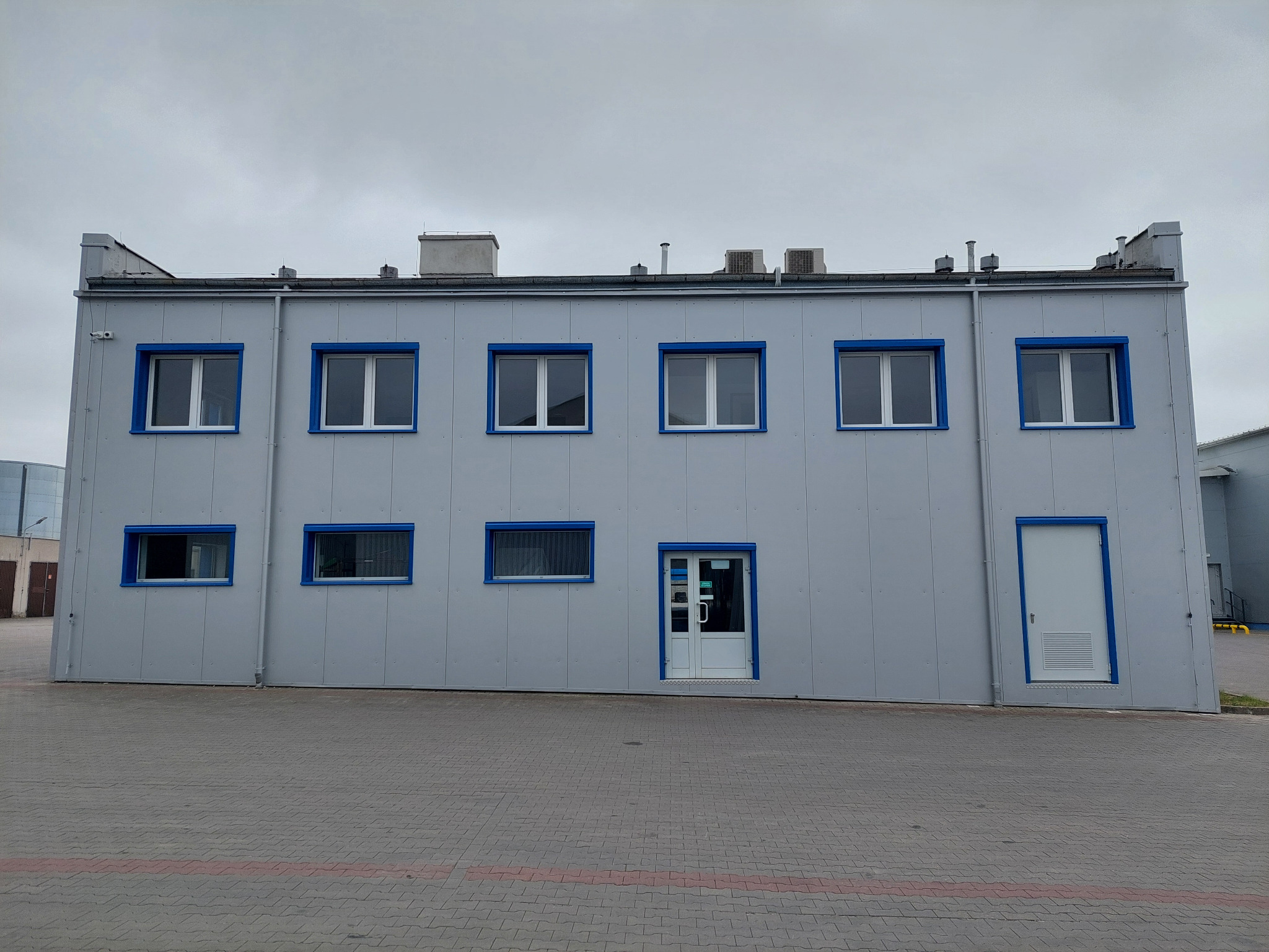 Dwukondygnacyjny budynek przemysłowy z szarych płyt warstwowych z niebieskimi obramowaniami okien i drzwi, widoczny z zewnątrz na tle pochmurnego nieba.
