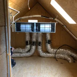 Centralna jednostka wentylacyjna z czterema elastycznymi przewodami w przestrzeni poddasza wykończonej płytami OSB, widoczne elementy instalacji elektrycznej i oświetlenie.