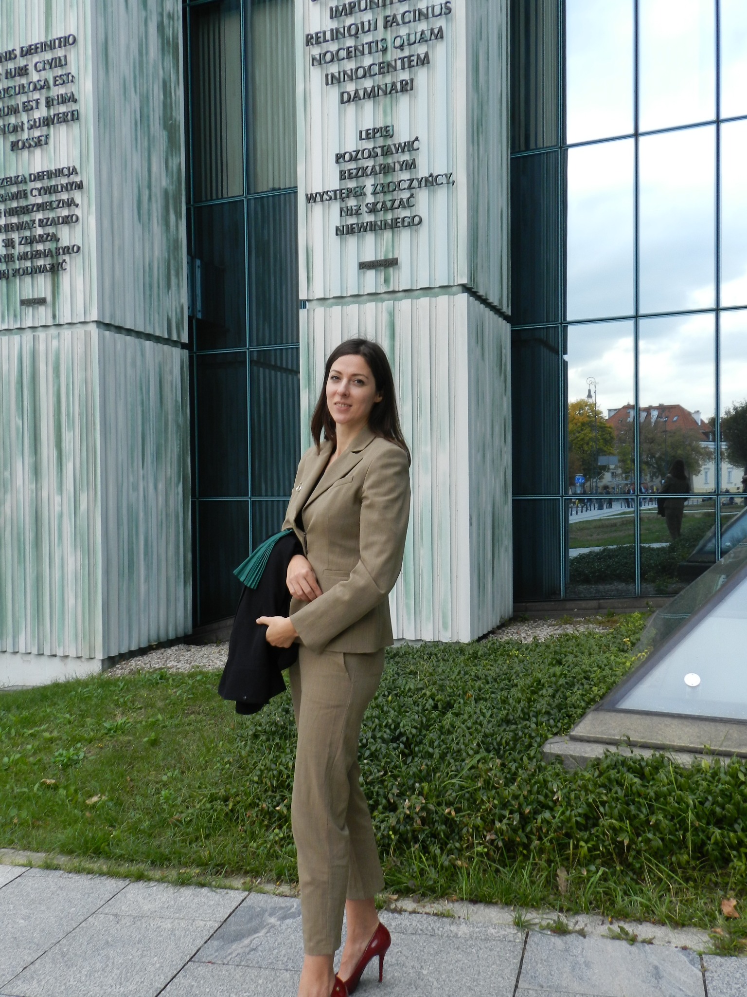 Kobieta w beżowym garniturze i czerwonych szpilkach stoi przed budynkiem sądu z inskrypcjami prawniczymi, trzymając togę.