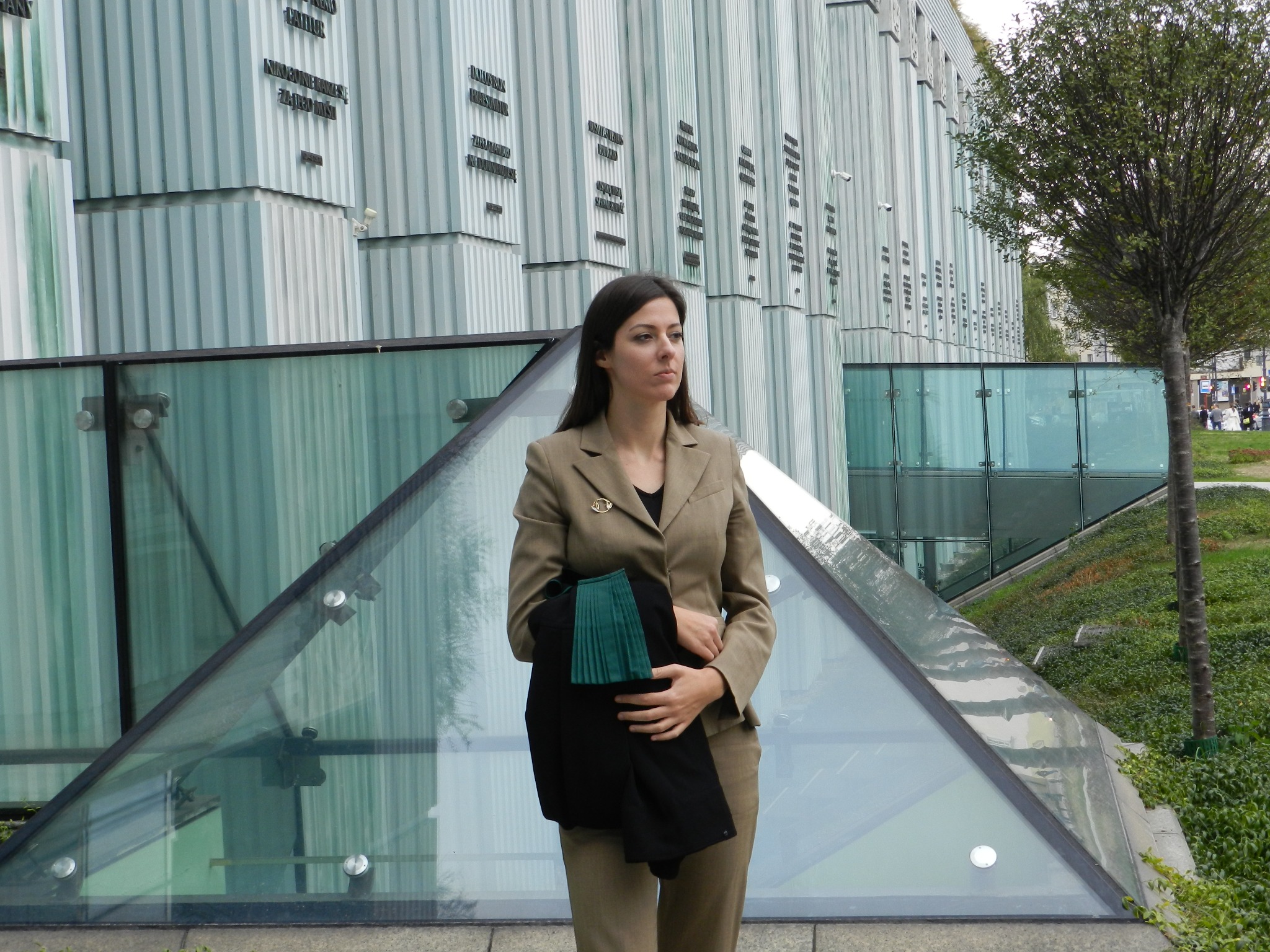 Kobieta w beżowym garniturze trzyma togę adwokacką na tle nowoczesnej architektury w Warszawie.