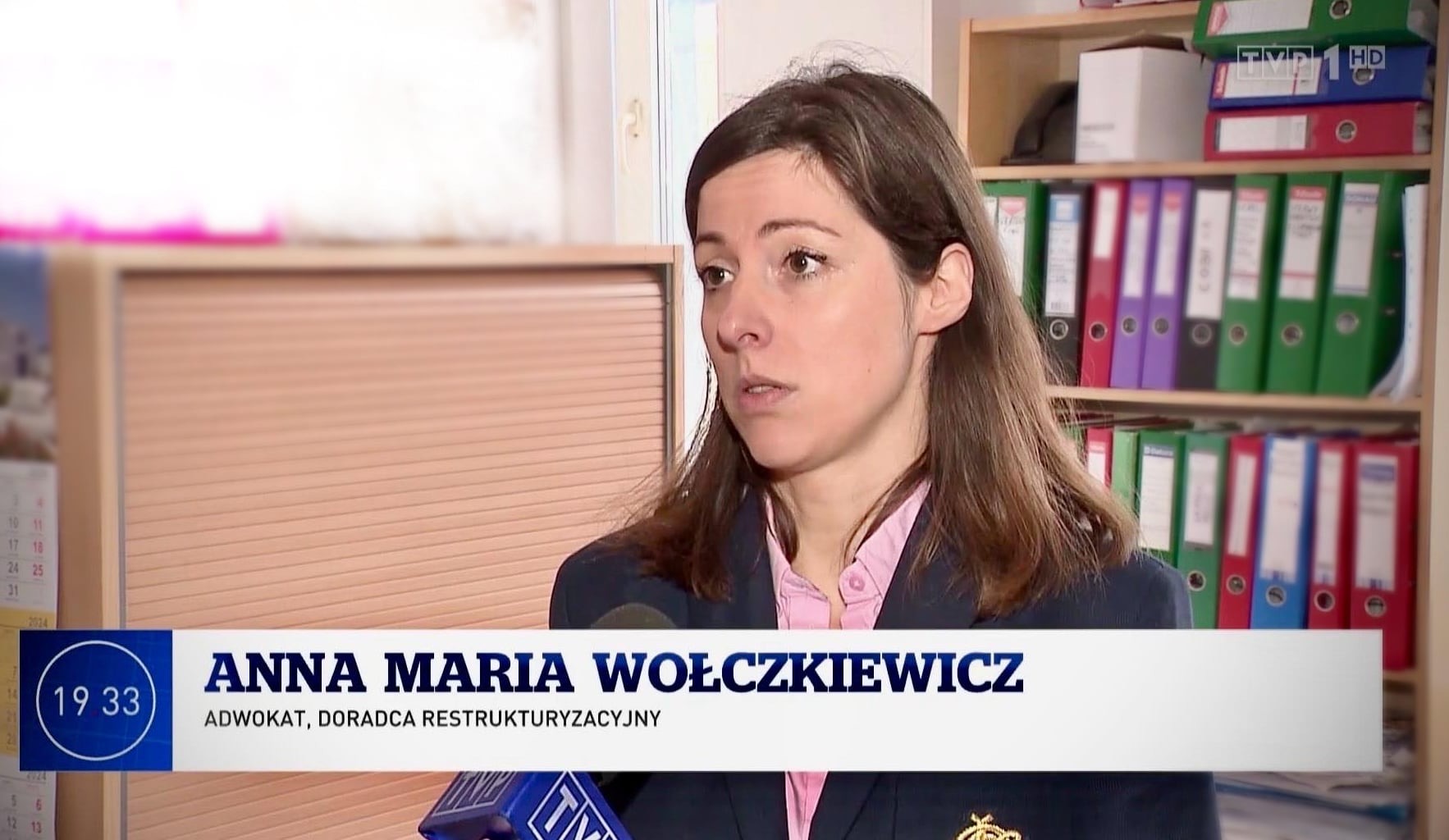 Kobieta w marynarce, prawdopodobnie podczas wywiadu telewizyjnego, na tle biura z roletą i regałem pełnym segregatorów.