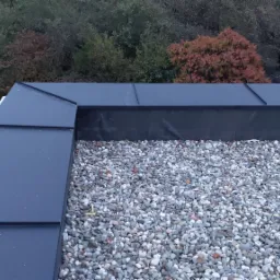 Wykończony dach płaski pokryty warstwą jasnych, okrągłych kamieni, z widocznymi obróbkami blacharskimi w kolorze grafitowym i fragmentem zieleni w tle.