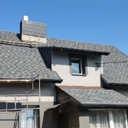 Dom jednorodzinny z szarym dachem z gontów bitumicznych w trakcie prac wykończeniowych, widoczne rusztowanie, częściowo obrobiony komin i zamontowane rynny.