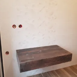 Drewniana, wisząca półka łazienkowa z ciemnego drewna na tle jasnych, strukturalnych płytek i drewnopodobnej podłogi; widoczne puszki elektryczne w ścianie.