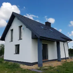 Gotowy, otynkowany dom jednorodzinny z ciemną dachówką i stolarką okienną, widok z zewnątrz w słoneczny dzień.