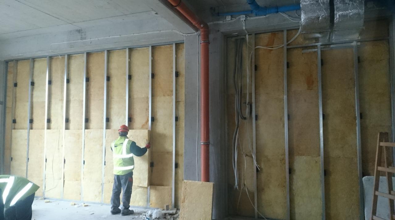 Prace izolacyjne ścian wewnętrznych wełną mineralną w metalowym stelażu. Widoczni pracownicy w odblaskowych kamizelkach. Pomarańczowa rura pionowa w tle.