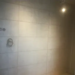 Ściana wyłożona jasnymi, matowymi płytami wielkoformatowymi z widocznymi puszkami instalacyjnymi i zwisającym przewodem elektrycznym, oświetlona punktowo.