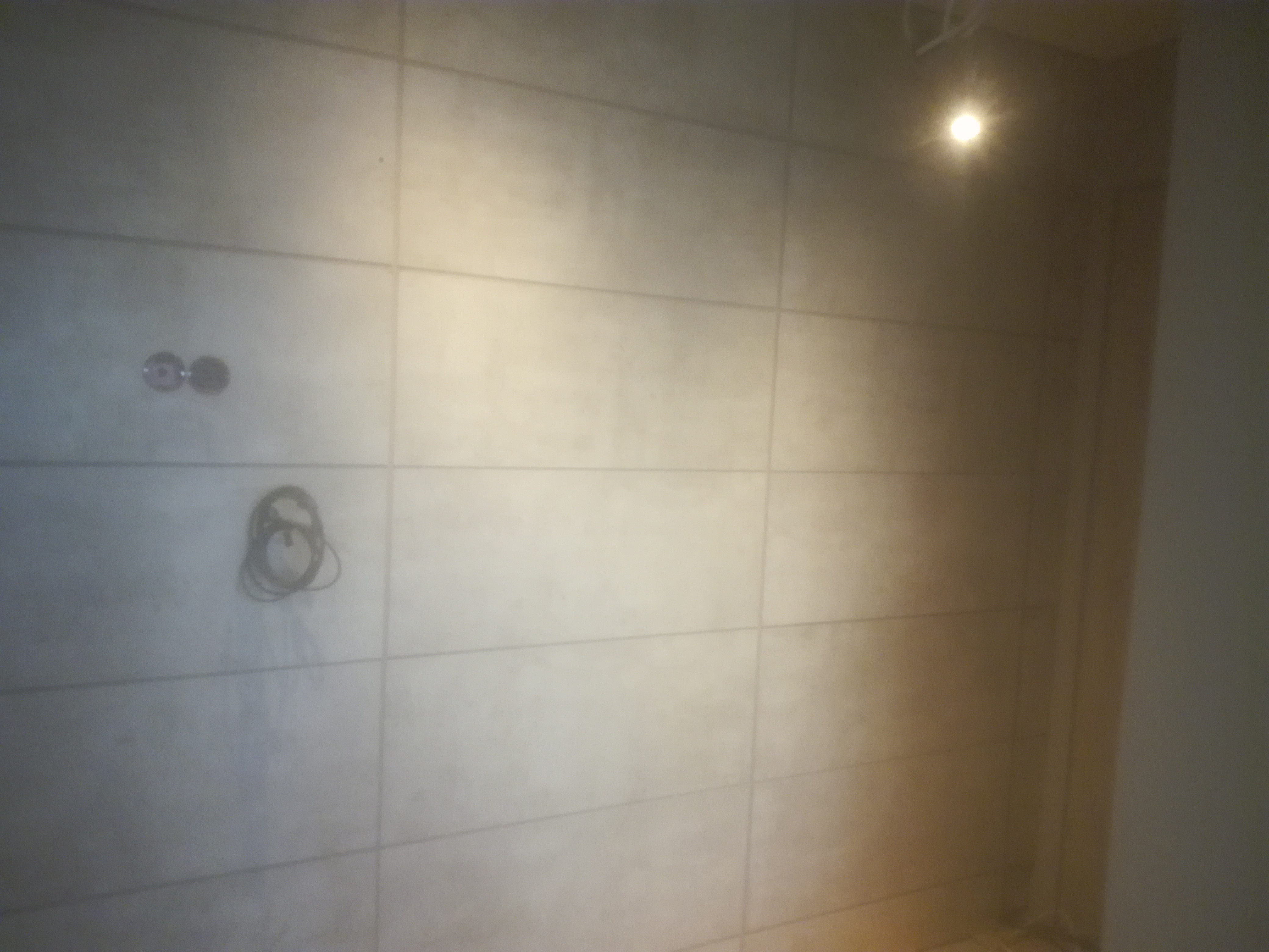 Ściana wyłożona jasnymi, matowymi płytami wielkoformatowymi z widocznymi puszkami instalacyjnymi i zwisającym przewodem elektrycznym, oświetlona punktowo.