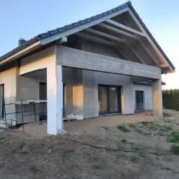 Dom w trakcie budowy z widoczną niezakończoną elewacją, betonowymi filarami werandy i przygotowanymi płytami styropianowymi.