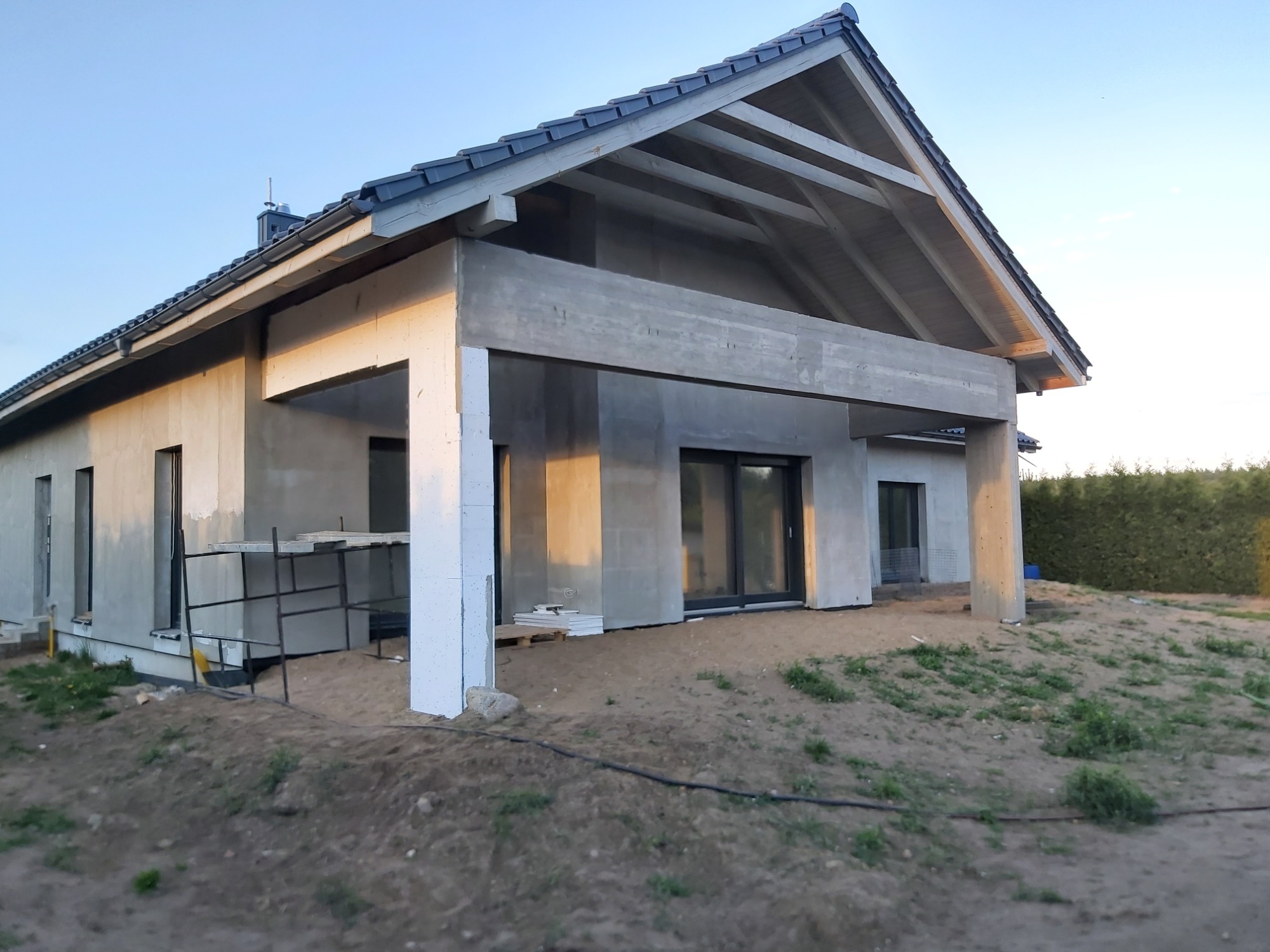 Dom w trakcie budowy z widoczną niezakończoną elewacją, betonowymi filarami werandy i przygotowanymi płytami styropianowymi.