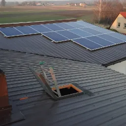 Panele słoneczne na brązowym dachu z blachy trapezowej, widoczna drabina prowadząca do włazu dachowego, w tle zabudowa jednorodzinna i pola uprawne w porannym świetle.