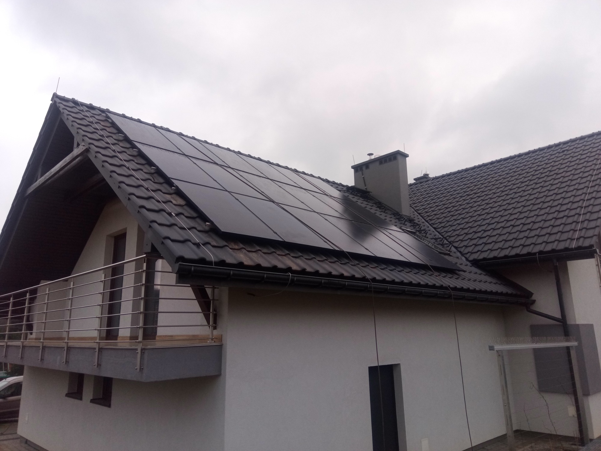 Panele słoneczne na dachu domu jednorodzinnego z balkonem, widoczne orynnowanie i komin, pochmurne niebo.