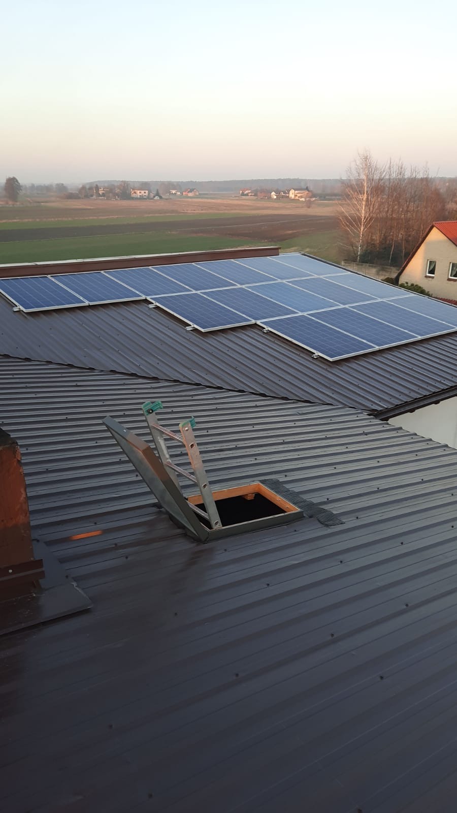 Panele słoneczne na brązowym dachu z blachy trapezowej, widoczna drabina prowadząca do włazu dachowego, w tle zabudowa jednorodzinna i pola uprawne w porannym świetle.