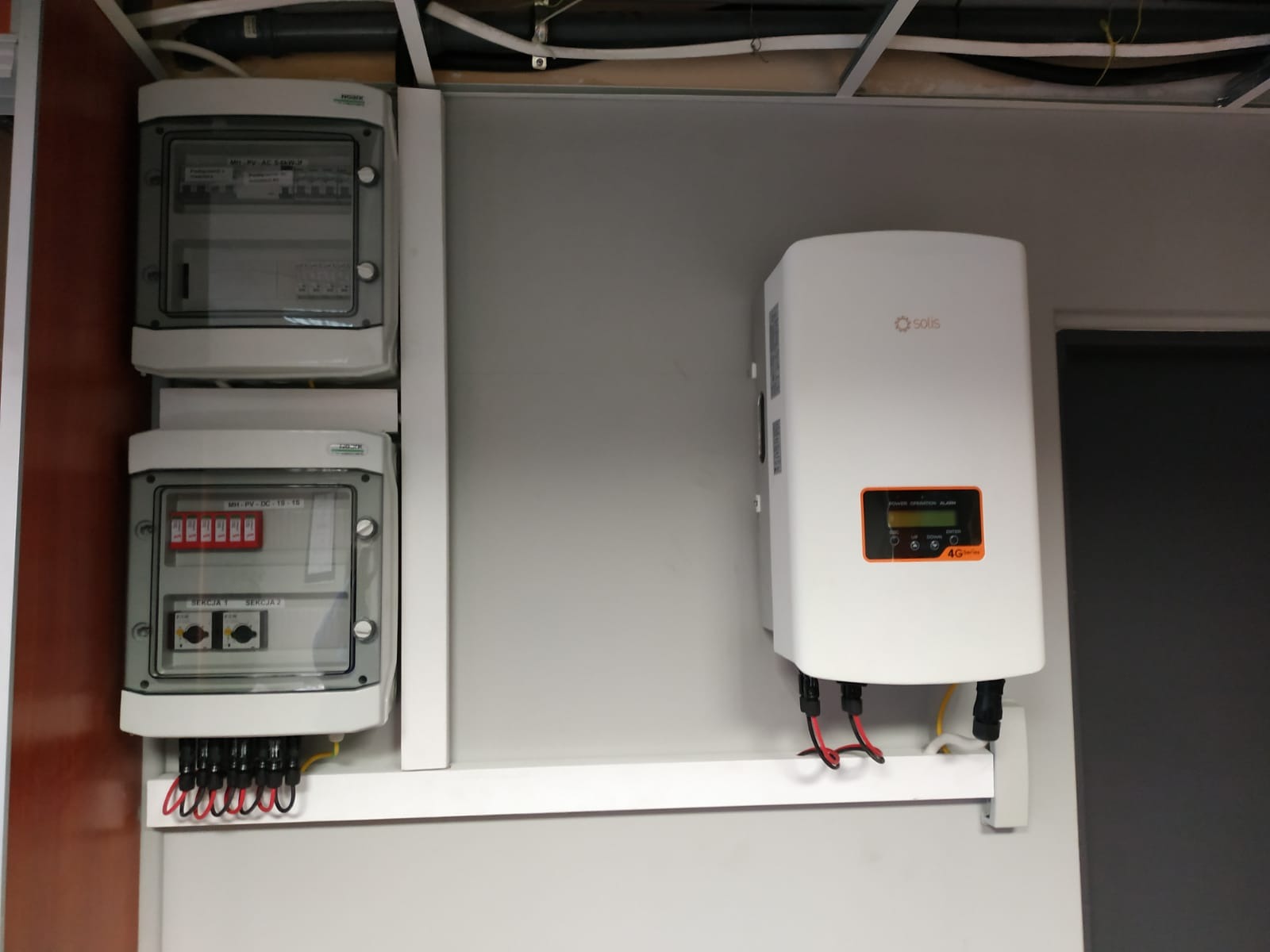Zamontowany biały falownik Solis z wyświetlaczem i zabezpieczenia elektryczne w dwóch szarych obudowach, okablowanie doprowadzone do urządzeń.