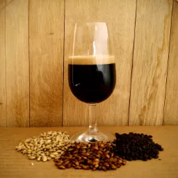 Kieliszek ciemnego piwa typu stout z jasną pianką, otoczony trzema rodzajami słodu (jasny, karmelowy i palony) na tle drewnianej beczki w Krakowie.