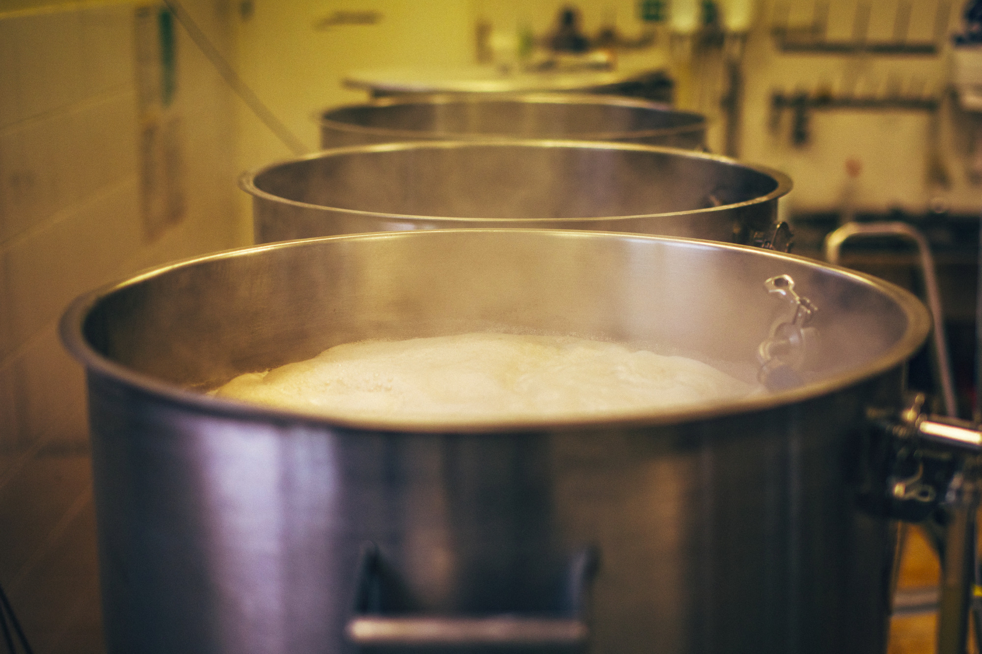 Rząd dużych, stalowych kadzi z gotującą się, pieniącą się zawartością, prawdopodobnie w browarze, z widocznymi elementami instalacji i wyposażenia w tle.