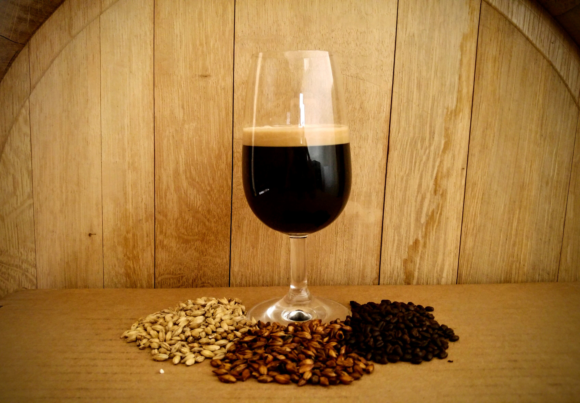Kieliszek ciemnego piwa typu stout z jasną pianką, otoczony trzema rodzajami słodu (jasny, karmelowy i palony) na tle drewnianej beczki w Krakowie.