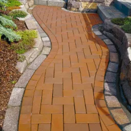 Kręta ścieżka z ceglastej kostki brukowej, obrzeża z szarego kamienia, rabaty z roślinami iglastymi i korą.