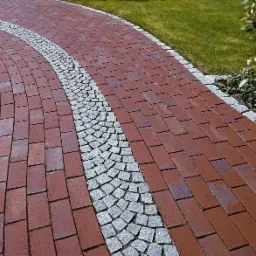 Czerwona ceglana ścieżka z dekoracyjnym pasem z szarej kostki granitowej, wijąca się przez trawnik i roślinność.