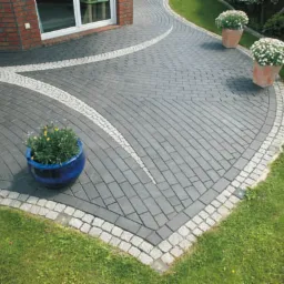 Ułożona nawierzchnia z szarej kostki brukowej z jasnymi obwódkami i wstawkami, donica z kwiatami w rogu, trawnik i fragment budynku w tle.