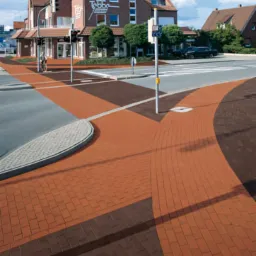 Ulica wyłożona kostką brukową w odcieniach brązu i czerwieni, tworząca wzór na skrzyżowaniu. W tle budynek z cegły i sygnalizacja świetlna.