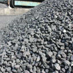 Kopalnia węgla: duży pryzmat bryłek węgla kamiennego, lekko wilgotnych i błyszczących, z fragmentem maszyny górniczej w tle.