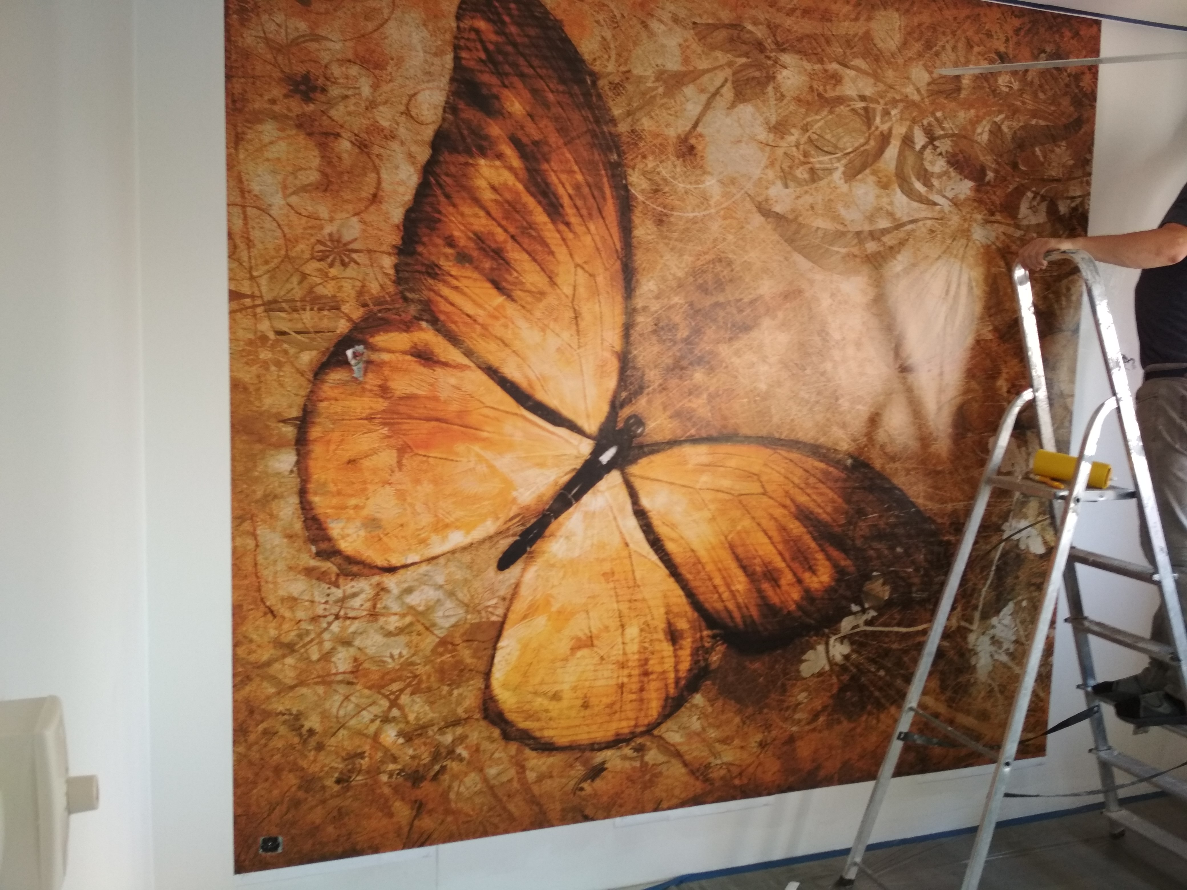 Naklejanie fototapety z motywem pomarańczowo-brązowego motyla na ścianie, widoczna drabina i fragment osoby montującej.