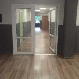 Korytarz z panelami podłogowymi w odcieniach brązu, ściany dwukolorowe: góra biała, dół szary, otwarte białe drzwi z widokiem na dalszą część korytarza i okno z zielenią na końcu.