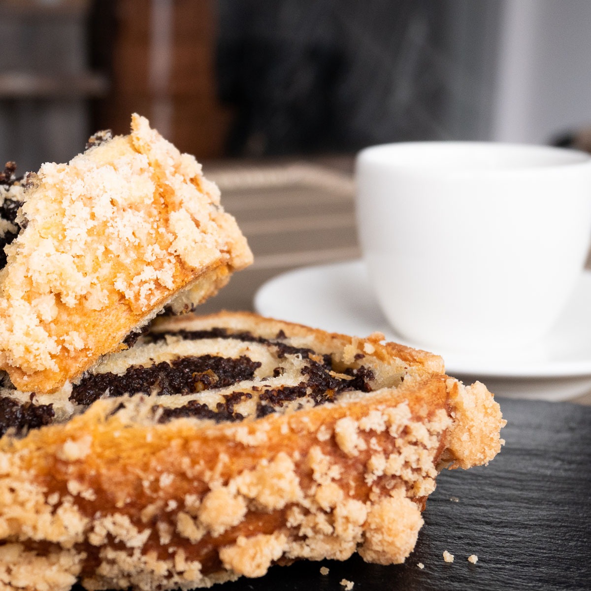 Dwa kawałki makowca z kruszonką na czarnym kamiennym talerzu, z filiżanką w tle, ukazujące warstwy maku i ciasta.