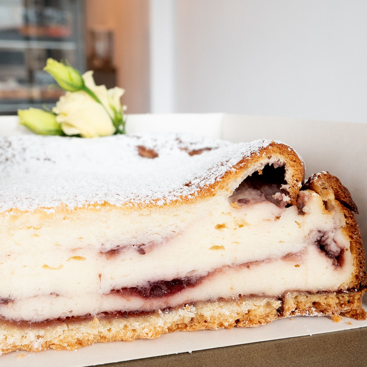 Kawałek sernika z owocami, posypany cukrem pudrem, z dekoracją z białych kwiatów w tle, umieszczony w białym kartonowym pudełku.