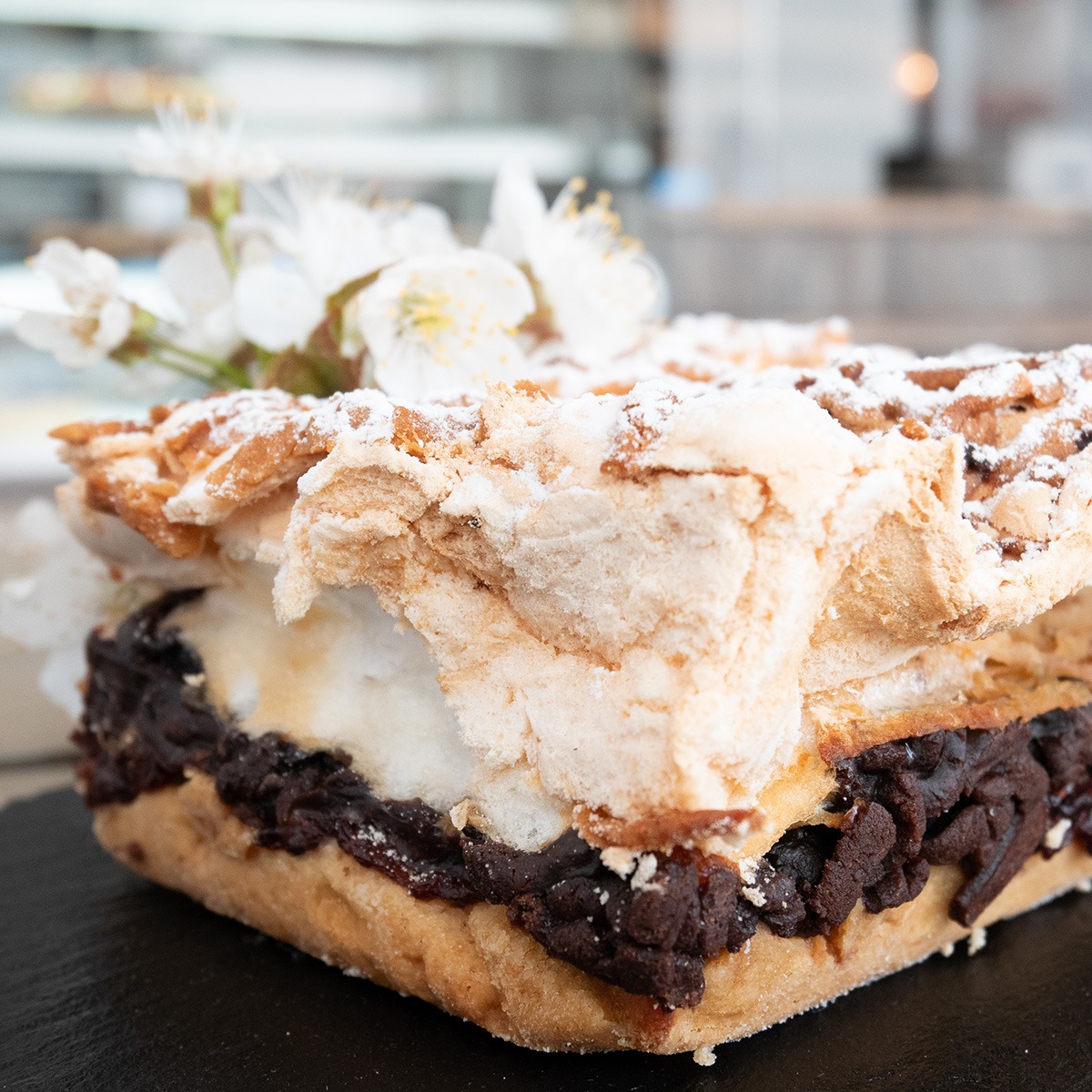 Kwadratowy kawałek pleśniaka z jabłkami, czarną porzeczką lub śliwkami, z bezową pianką i kruszonką, udekorowany gałązką kwitnącej wiśni, na czarnym talerzu.