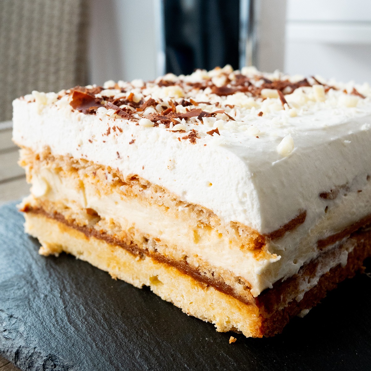 Kwadratowy kawałek ciasta 3-bit na czarnym, kamiennym talerzu. Widoczne warstwy: biszkopt, masa krówkowa, krem budyniowy i bita śmietana posypana startą czekoladą.