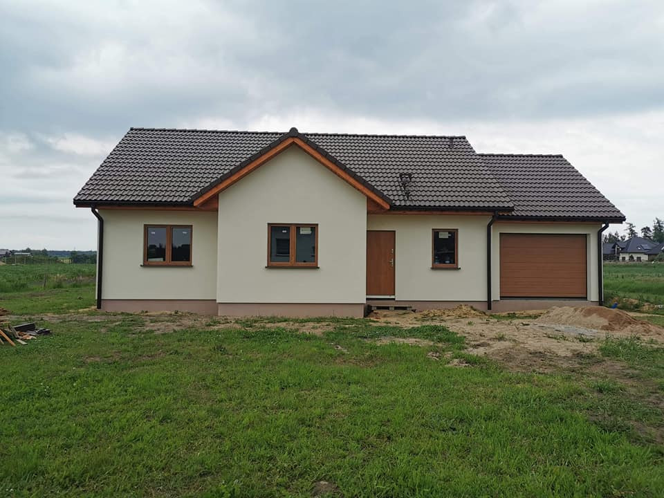 Parterowy dom z garażem, kremowa elewacja, brązowa dachówka i stolarka okienna, widok z przodu na trawniku.