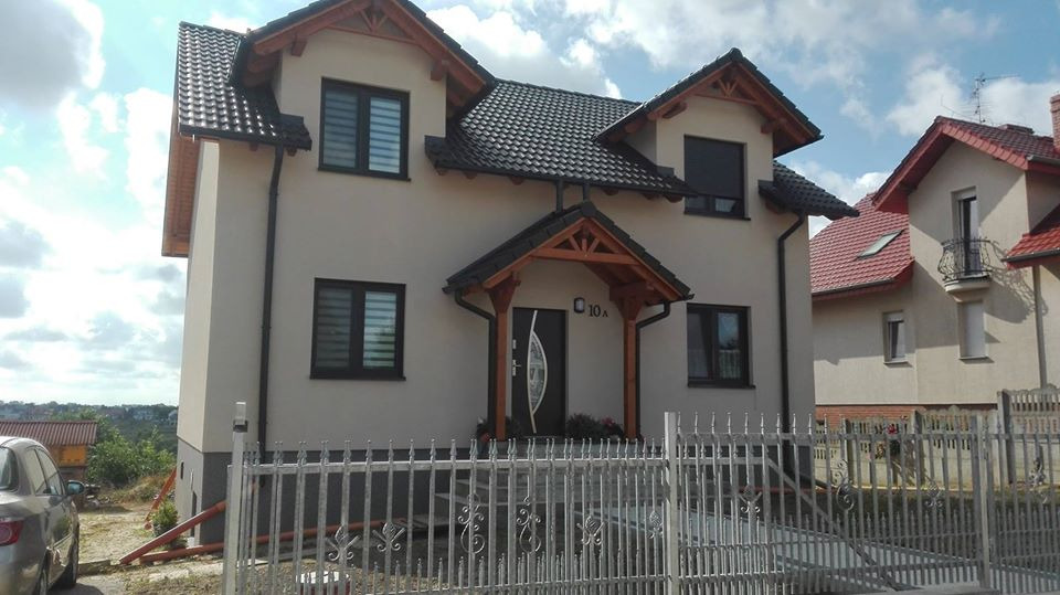 Nowoczesny dom jednorodzinny z ciemną dachówką i drewnianym podcieniem wejściowym, ogrodzony metalowym płotem w Ostrowie Wielkopolskim.