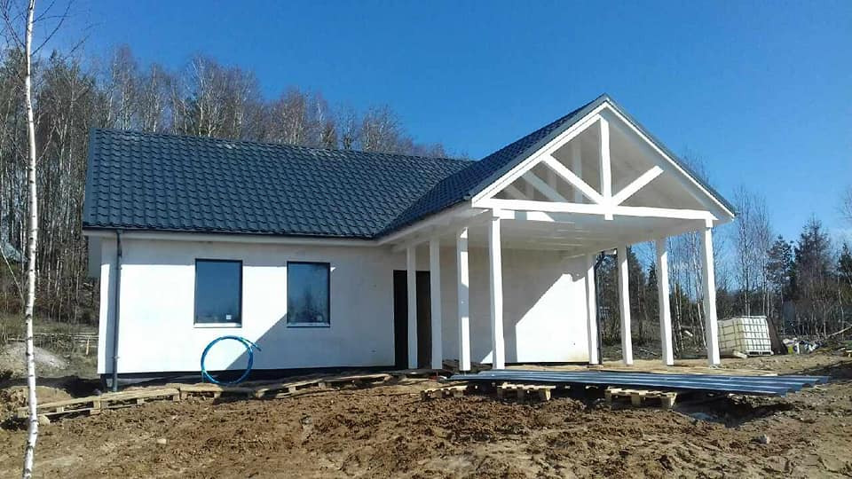 Niedokończony biały budynek z ciemnym dachem i zadaszeniem wspartym na kolumnach, widoczne palety i rury na ziemi, w tle drzewa.