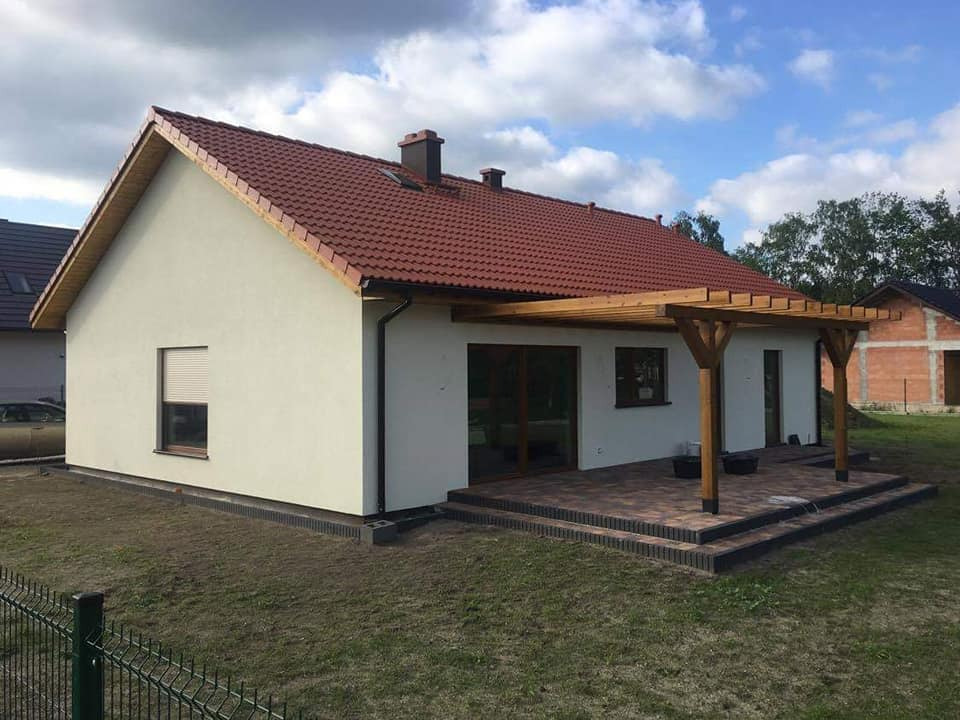 Niski parterowy dom z jasną elewacją, czerwoną dachówką i drewnianą pergolą z tarasem wyłożonym kostką, widoczne obrzeża z ciemnej kostki, trawnik i ogrodzenie.