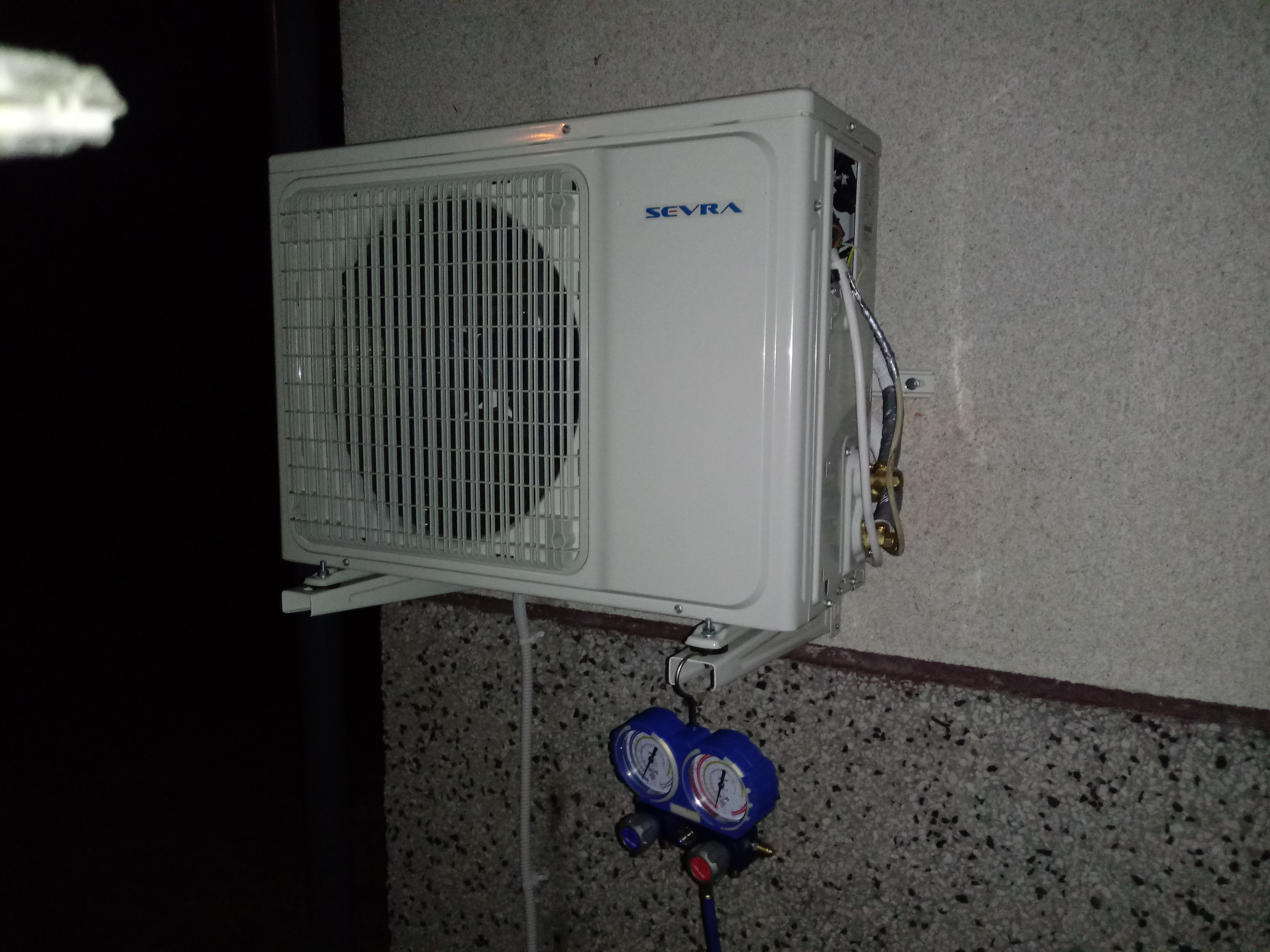 Biała jednostka zewnętrzna klimatyzacji marki Sevra zamontowana na szarej, chropowatej ścianie; pod nią podwieszony manometr serwisowy z niebieską obudową.