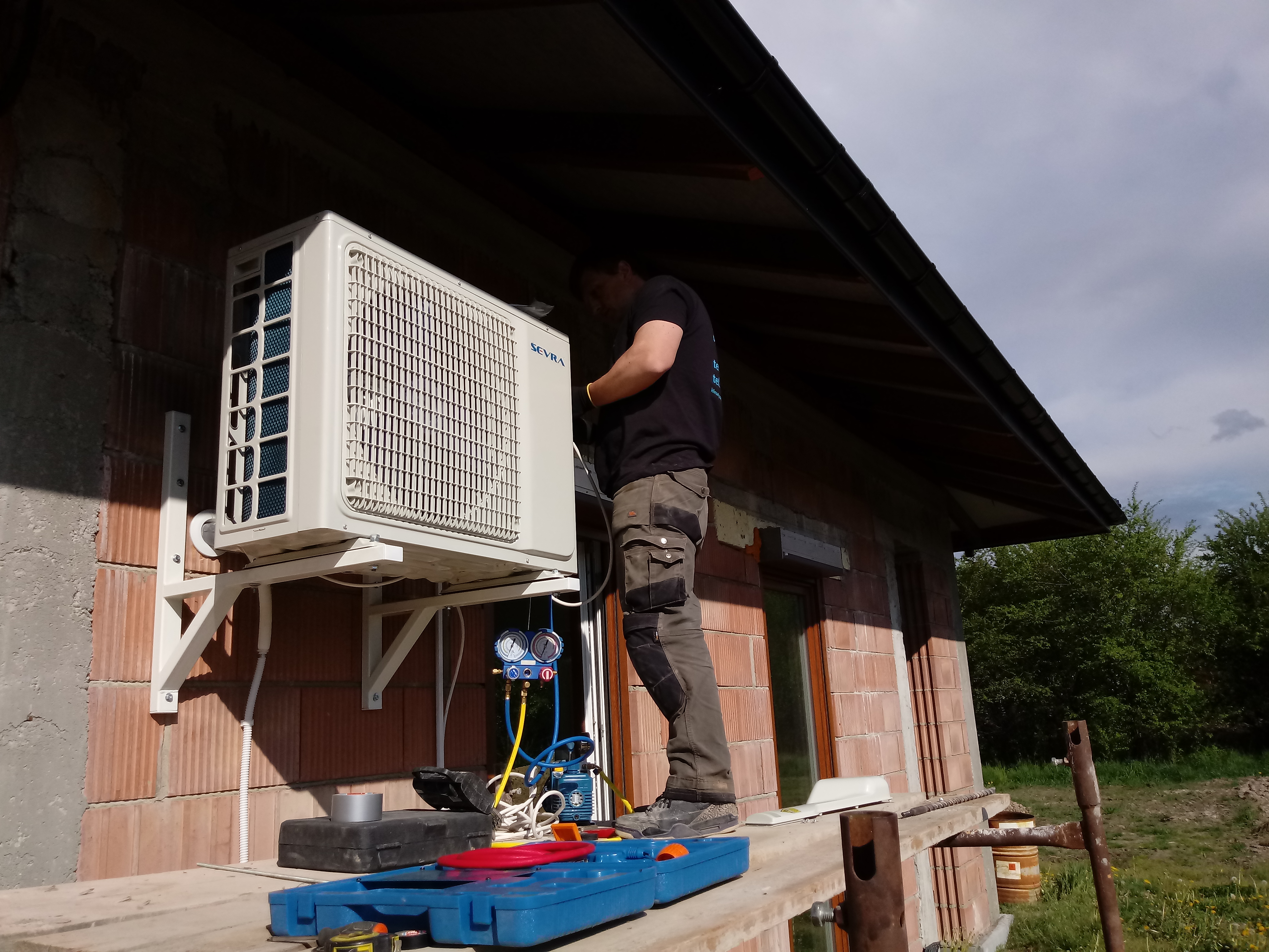 Instalator w czarnej koszulce i roboczych spodniach, montuje jednostkę zewnętrzną klimatyzatora marki Sevra na ceglanej ścianie domu, używając narzędzi pomiarowych i skrzynki narzędziowej.